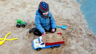 Küçük bir çocuk kum havuzunda oynarken oyuncak bir kamyonu kumla doldururken küçük bir traktör yakınlarda oturuyor. Güneş bu neşeli açık havada parlıyor..