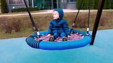 Mavi paltolu sıcacık giyinmiş genç bir çocuk canlı bir parkta büyük yuvarlak bir salıncakta oturuyor. Çevre bereketli, huzurlu bir atmosfer sergiliyor..
