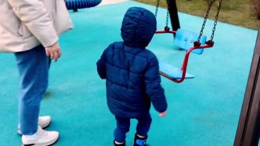 Mavi ceketli bir çocuk salıncağa doğru heyecanlı bir şekilde hareket ederek oyun alanını keşfediyor. Yetişkin biri bu keyifli gezide güvenliği sağlamak için nöbet tutuyor..