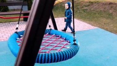Mavi ceketli küçük bir çocuk oyun alanında eğlenirken büyük bir salıncağa doğru yürüyor. Bölgede canlı yüzeyler ve eğlenceli yapılar yer alıyor.