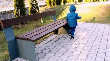 Mavi ceket giyen ve eşleşen pantolon giyen genç bir çocuk ahşap bir bankın yanındaki kaldırımlı yolda yürüyor. Güneş, sakin bir park batımında ışıldıyor..
