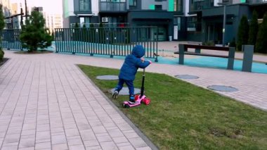 Mavi ceketli küçük bir çocuk, şehir merkezinde asfalt bir yol boyunca pembe bir scooter kullanıyor. Çocuğun serin bir günde dışarıda oyun oynamaktan hoşlanması gibi atmosfer de hoş..