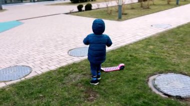 Sıcak kıyafetler giymiş küçük bir çocuk parktaki kaldırımlı bir patika boyunca yürür, yakınlardaki çimlerin üzerinde park etmiş pembe bir scooter 'a ilgi gösterir..