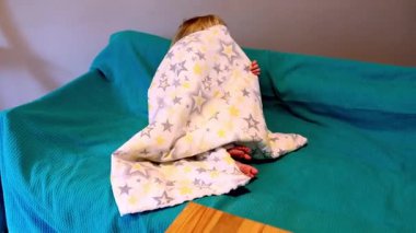 Neşeli bir çocuk, yıldızlarla kaplı yumuşak bir battaniyeye sarılmış bir kanepede oturur. Kahkahalar oynarlarken atmosferi doldurur, mutluluk ve rahatlığı kapalı bir ortamda gösterir..