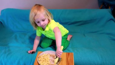 Parlak yeşil elbiseli genç kanepeye oturur ve tepsiden yemek yerken mutlu olur. Çocuk rahat bir ev atmosferiyle çevriliyken yiyeceğe odaklanıyor..