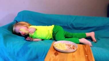 Parlak giyinmiş genç bir çocuk, mavi bir kanepede uzanmış, yakındaki bir masaya yerleştirilmiş abur cuburlara uzanıyor. Odanın neşeli bir atmosferi var..