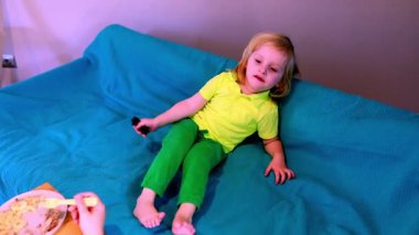 Parlak yeşil giyinmiş genç bir çocuk mavi bir battaniyeye rahatça oturur. Rahat ortam, kapalı oyun zamanı boyunca sakin bir anı gösteren yakınlardaki abur cuburları içeriyor.