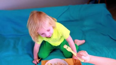 Küçük bir çocuk kanepede oturup çatalla tabaktan yemeğin tadını çıkarıyor. Kıyafetin ve kanepenin parlak renkleri neşeli bir atmosfer yaratır..