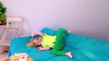 Parlak sarı gömlekli ve yeşil pantolonlu bir çocuk mavi bir kanepede zıplıyor. Neşeli atmosfer, rahat ve renkli bir yaşam alanındaki çocukların enerjisini ve heyecanını vurgular..