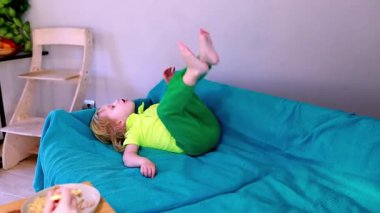 Küçük bir çocuk, güneşli bir odadaki mavi bir kanepede, meyve ve atıştırmalıklarla dolu bir masa ile rahatça uzanıyor..