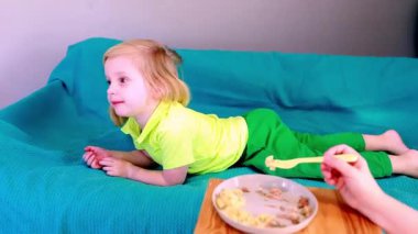 Küçük bir çocuk yemek yerken kanepede yatarken eğlenir. Çevre rahatlatıcıdır ve çocuk yemekle etkileşim halindeyken merak ve haz gösterir..