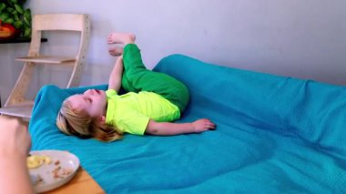 Küçük çocuk mavi bir kanepede gevşiyor, bir tabak yemek yerken eğlenceli aktivitelerle meşgul oluyor. Oda canlı ve bitkilerle dolu..
