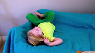 Küçük bir çocuk mavi bir kanepede rahat bir şekilde uzanır. Renkli giysiler içinde, bir anlık rahatlığın tadını çıkarırken. Ev atmosferi neşeli havayı arttırır..