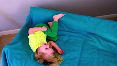 Genç bir çocuk neşeyle yuvarlanır ve yuvarlanır parlak mavi bir kanepede, neşeli hareketler sergiler ve kaygısız bir ruh kapalı bir ortamda.