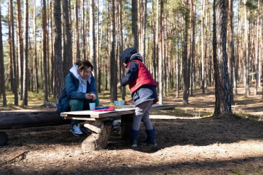 Ormandaki bir bankta oturan bir kadın telefonunu kontrol ederken, kırmızı yelekli bir çocuk da yanında küçük bardaklarla oynuyor. Uzun ağaçlar etraflarını sarar ve huzurlu bir atmosfer yaratır..