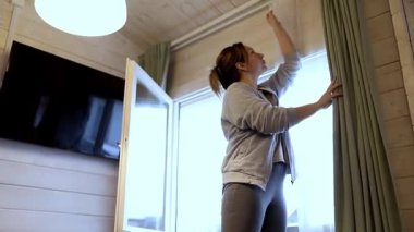 Kırsal bir odada duran bir kadın, doğal ışık için yeşil perdeleri açıyor. Uzay, ahşap duvarlar ve sıcak bir atmosfer yaratan bir televizyon barındırıyor..