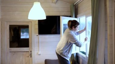 Bir kadın perdeleri açıyor ve rahat bir kulübenin camlarını temizliyor. Güneş ışığı akarak uzayın sıcak atmosferini arttırır. Parlak bir öğleden sonra..