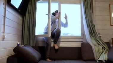 Bir kadın sıcak ve modern bir evin penceresine ulaşmak ve temizlemek için kanepeye tırmanıyor. Güneş ışığı odayı doldurur, taze ve misafirperver atmosferi güçlendirir..
