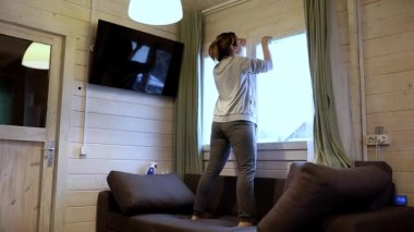 Bir kadın kanepede duruyor, büyüleyici bir kamaradaki perdeleri düzeltiyor. Uzay, ahşap duvarlar ve modern bir tasarım içeriyor. Rahatlamak için sıcak bir atmosfer sağlıyor..