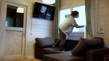 Bir kadın kanepede durur, şık ahşap bir kulübede perdeleri ayarlamak için uzanır. Doğal ışık odayı dolduruyor ve uzayın davetkar atmosferini arttırıyor..