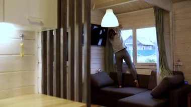 Sıcak ve davetkar bir kamaranın oturma odasındaki pencere perdelerini indirmek için bir kişi kanepede durur. Doğal ışık boşluğu doldurur, ahşap detayları ve konforlu mobilyaları vurgular..