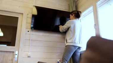 Bir kadın, sıcak ve davetkar bir oturma odasının duvarına şık bir televizyon monte ediyor. Kurulumu tamamlarken güneş ışığı büyük pencerelerden süzülür.