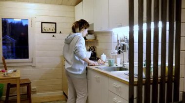 Rahat giyinmiş bir kadın sıcak bir mutfakta yemek hazırlıyor. Sıcak ışık, tezgahtaki malzemeleri düzenlerken hoş bir atmosfer yaratıyor..