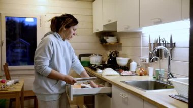 Bir kadın sıcak bir mutfakta yemek hazırlarken çekmeceden malzemeleri düzenliyor. Ilık ışık, akşam yemek yaparken ev gibi bir atmosfer yaratıyor..