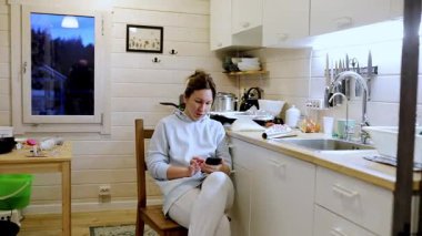 Bir kadın, yemek pişirme aletleri ve doğal ışıkla çevriliyken, rahat bir mutfakta, telefonuna dalmış bir şekilde rahat bir şekilde oturur. Ortam huzurlu ve davetkar..