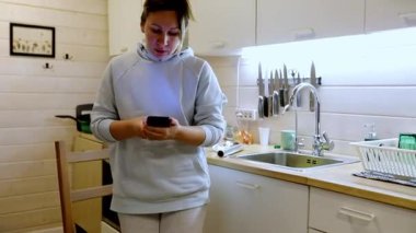 Rahat kıyafetler giymiş, modern bir mutfakta rahat bir kadın sandalyeye oturmuş ve akıllı telefonuyla meşgul oluyor. Hava sıcak ve davetkar..