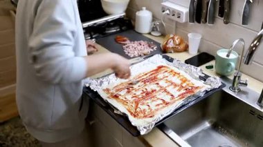 Bir insan mutfakta hamurun üzerine sos döker. Doğranmış etler ve diğer malzemeler yakınlarda, fırında pişirmeden önce montaj için hazırlandı..