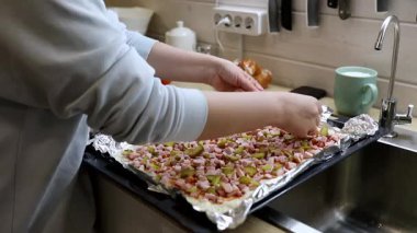 Elleri ustalıkla doğranmış sebzeleri ve eti, mutfak odasındaki folyoyla kaplı bir kabın üzerine koyuyor. Taze malzemeler sergileniyor, ev yapımı yemek atmosferini yansıtıyor..