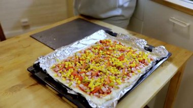 Bir insan mutfakta canlı bir sebze pizzası yapıyor. Üste çeşitli taze malzemeler yayılıyor, yaratıcı bir pişirme sürecini vurguluyorlar..