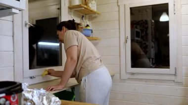 Sıcak, davetkar bir mutfakta, bir kadın gelecek güne hazırlanırken bir süngerle yüzeyleri özenle siler. Yumuşak ışık sıcak atmosferi güçlendirir.