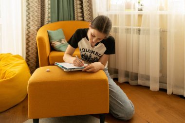 Genç bir kız yerde oturur ve defterine yazmaya konsantre olur. Parlak oturma odasında turuncu bir kanepe ve renkli yastıklar var. Sıcak bir atmosfer yaratıyorlar..