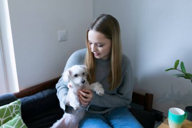 Uzun saçlı bir kadın kanepeye oturur ve kucağında küçük bir köpek beşiği taşır. Oturma odasında yumuşak ışıklandırma ve basit bir dekor var. Sıcak bir atmosfer yaratıyor..