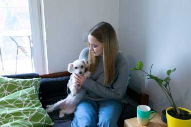 Genç bir kadın kanepeye oturmuş, kucağında küçük bir köpek, aydınlık ve modern bir oturma odasında huzurlu bir anın tadını çıkarıyor. Yakınlarda, bir kahve fincanı ve bitkiler sıcak atmosfere eklenir..