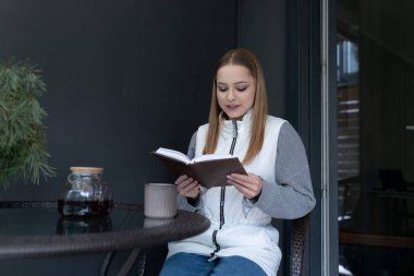 Genç bir kadın şık bir terasta rahatça oturup kitap okuyor ve kahvesinin tadını çıkarıyor. Atmosfer huzurlu ve davetkar, rahatlamak için mükemmel..