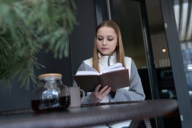 Genç bir kadın şık bir kafede, yanında bir fincan sıcak kahveyle kitap okumaya dalmış bir şekilde bir masada oturuyor. Ortam sakin, sakin dakikalar için mükemmel..