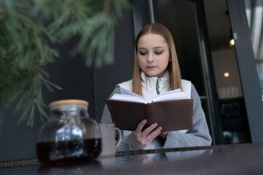Genç bir kadın, sıcak bir içecekten yudumlarken bir kitaba dalmış modern bir kahve dükkanında oturuyor. Davetkar atmosfer rahatlamayı ve odaklanmayı teşvik eder..