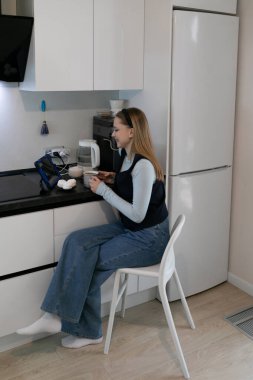 Çağdaş bir mutfakta sandalyeye oturmuş sıcak bir şeyler içip hafif bir şeyler atıştıran bir kadın. Uzay, lüks dolaplar ve aletler içeriyor, rahat bir atmosfer yaratıyor..