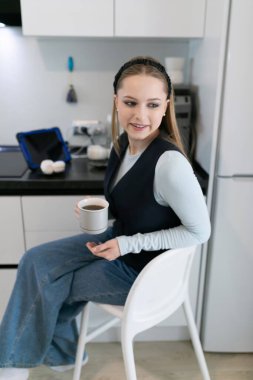 Genç bir kadın, gösterişli bir mutfakta beyaz bir sandalyede, elinde bir fincan kahveyle rahatça oturuyor. Günlük kıyafetinde açık mavi bir bluz ve geniş bacaklı kot pantolon var. Rahat bir hava yaratıyor..