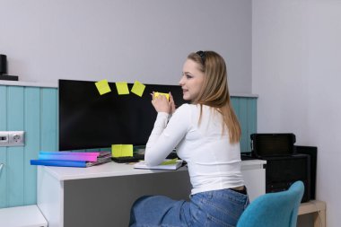 Genç bir kadın rahat bir ofis masasında oturmuş notları gözden geçiriyor ve bir monitörde renkli yapışkan notlarla çevrili. Güneş ışığı içeri girer ve sıcak bir atmosfer yaratır..