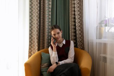 Şık giyinmiş genç bir kadın canlı sarı bir koltukta otururken telefonla konuşuyor. Parlak perdeler ve dekoratif yastıklar rahat atmosferi güçlendirir.