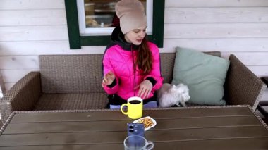 Parlak pembe ceketli genç bir kadın kahve ve atıştırmalıklarla bir masada oturuyor. Küçük beyaz köpeği yanında dinlenirken o atmosferin tadını çıkarıyor..