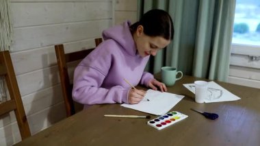 Genç bir kadın, çizim ve resim yaparak evde huzurlu bir öğleden sonranın tadını çıkarıyor. Ahşap bir masaya oturmuş, sanat eserlerine odaklanmış, etrafını sanat malzemeleri ve sıcak bir içecekle sarmış..