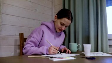 Sıcak bir kulübede, genç bir sanatçı sıcak bir içeceğin tadını çıkarırken yaratıcı fikirlerini hayata geçirmek için fırça ve boya kullanarak çizimlerine dikkatle odaklanır..