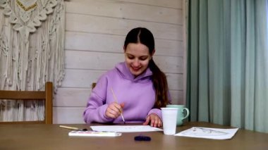 Genç bir sanatçı ahşap bir masada otururken suluboya resmine odaklanır. Yumuşak ışık çalışma alanını aydınlatıyor, yaratıcılık sürecini geliştiriyor..