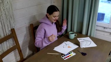 Genç bir kadın ahşap bir masada suluboya resimlerle oturuyor. Gazeteler, fırçalar ve bir fincan çayla çevrili bir kulübede sakin bir öğleden sonranın tadını çıkarıyor. Yaratıcı enerji havayı doldurur..