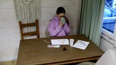 Sıcak bir odadaki ahşap bir masada oturan bir insan, etrafını boyama malzemeleriyle sarmışken bardaktan yudumluyor. Masada renkli sanat eserleri sergileniyor ve sakin bir atmosfer yaratıyor..